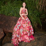 A model wearing a strapless pink floral ball gown stands on rocks surrounded by tall green plants at night.