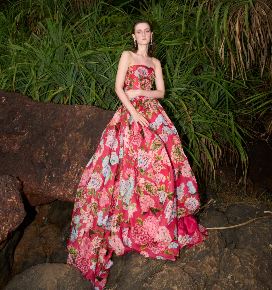 A model wearing a strapless pink floral ball gown stands on rocks surrounded by tall green plants at night.