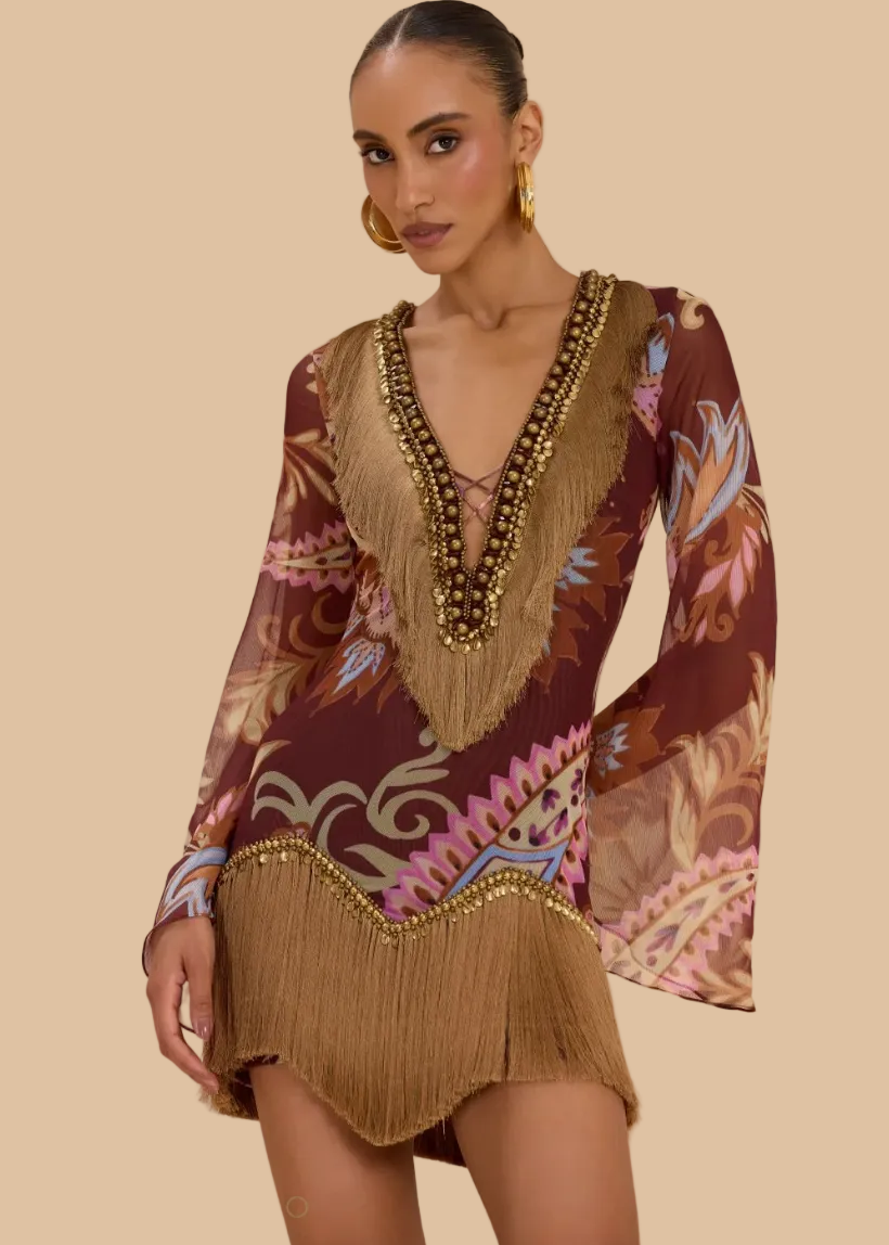 A woman models a bohemian-style mini dress with a brown base, multicolor floral and scroll patterns, and prominent tan fringe detailing along the neckline, sleeves, and hem. She is wearing gold hoop earrings and a chunky gold beaded necklace against a soft beige studio backdrop.