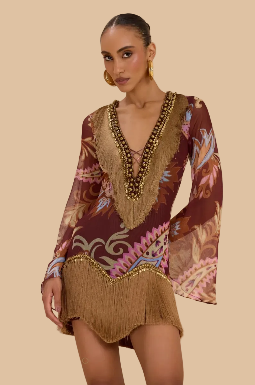 A woman models a bohemian-style mini dress with a brown base, multicolor floral and scroll patterns, and prominent tan fringe detailing along the neckline, sleeves, and hem. She is wearing gold hoop earrings and a chunky gold beaded necklace against a soft beige studio backdrop.