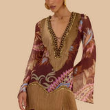 A woman models a bohemian-style mini dress with a brown base, multicolor floral and scroll patterns, and prominent tan fringe detailing along the neckline, sleeves, and hem. She is wearing gold hoop earrings and a chunky gold beaded necklace against a soft beige studio backdrop.