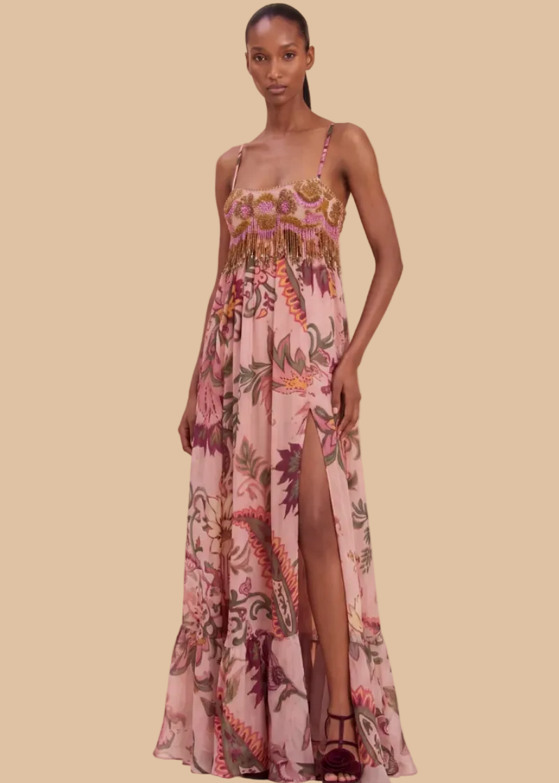 A model wears a floor-length pink floral dress with thin straps, a smocked bodice, and a high leg slit, standing against a soft beige draped backdrop.