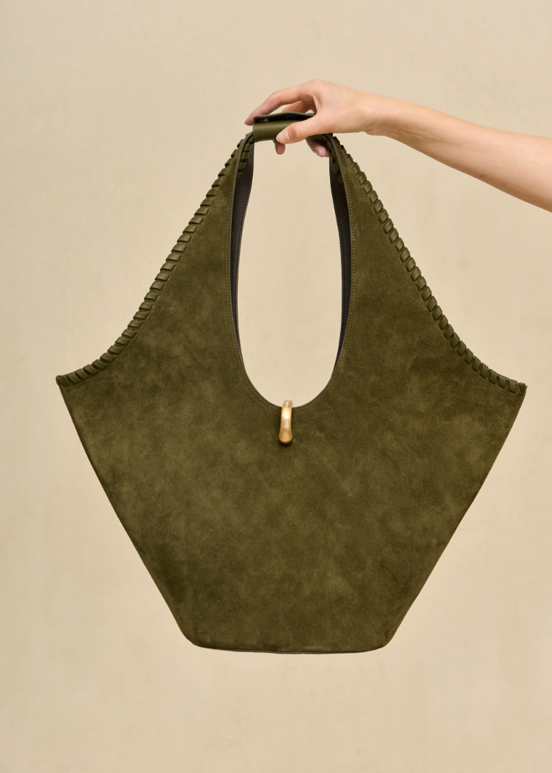 An olive green suede shoulder bag with a wide top and braided trim, held by a person's hand against a plain beige background.
