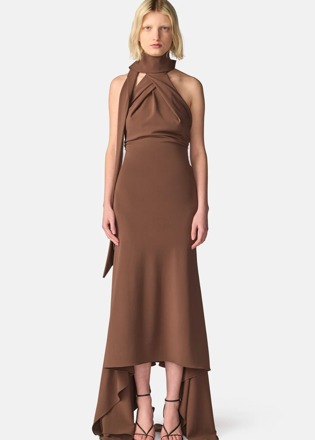 A model wearing a high-neck, halter-style brown dress with a draped side detail and a high-low hemline, paired with black strappy heels, against a light gray studio background.