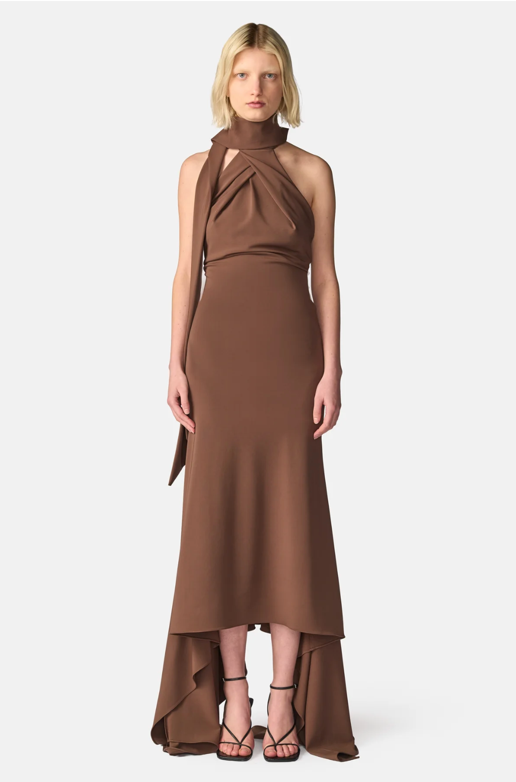 A model wearing a high-neck, halter-style brown dress with a draped side detail and a high-low hemline, paired with black strappy heels, against a light gray studio background.