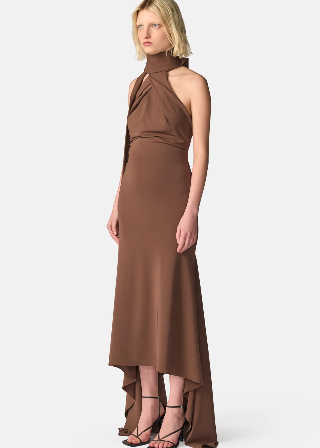 A model wearing a high-neck, halter-style brown midi dress with a draped, gathered bodice and an asymmetrical high-low hemline, paired with black strappy heels, against a light gray studio background.