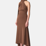A model wearing a high-neck, halter-style brown midi dress with a draped, gathered bodice and an asymmetrical high-low hemline, paired with black strappy heels, against a light gray studio background.