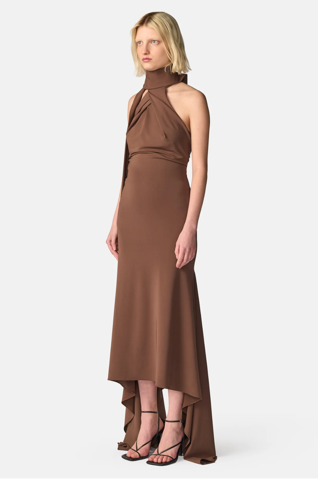 A model wearing a high-neck, halter-style brown midi dress with a draped, gathered bodice and an asymmetrical high-low hemline, paired with black strappy heels, against a light gray studio background.