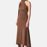 A model wearing a high-neck, halter-style brown midi dress with a draped, gathered bodice and an asymmetrical high-low hemline, paired with black strappy heels, against a light gray studio background.