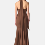 A woman viewed from the back wearing a floor-length brown halter gown with a draped wrap detail down the back, paired with black strappy heels, against a light gray studio background.