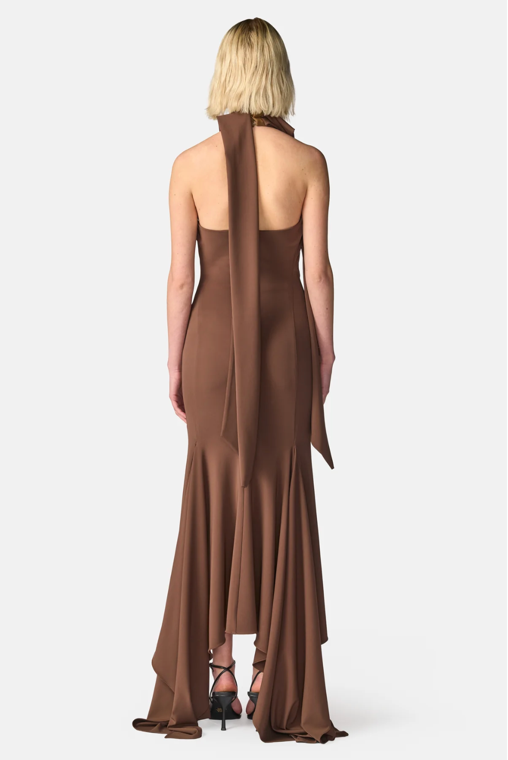A woman viewed from the back wearing a floor-length brown halter gown with a draped wrap detail down the back, paired with black strappy heels, against a light gray studio background.
