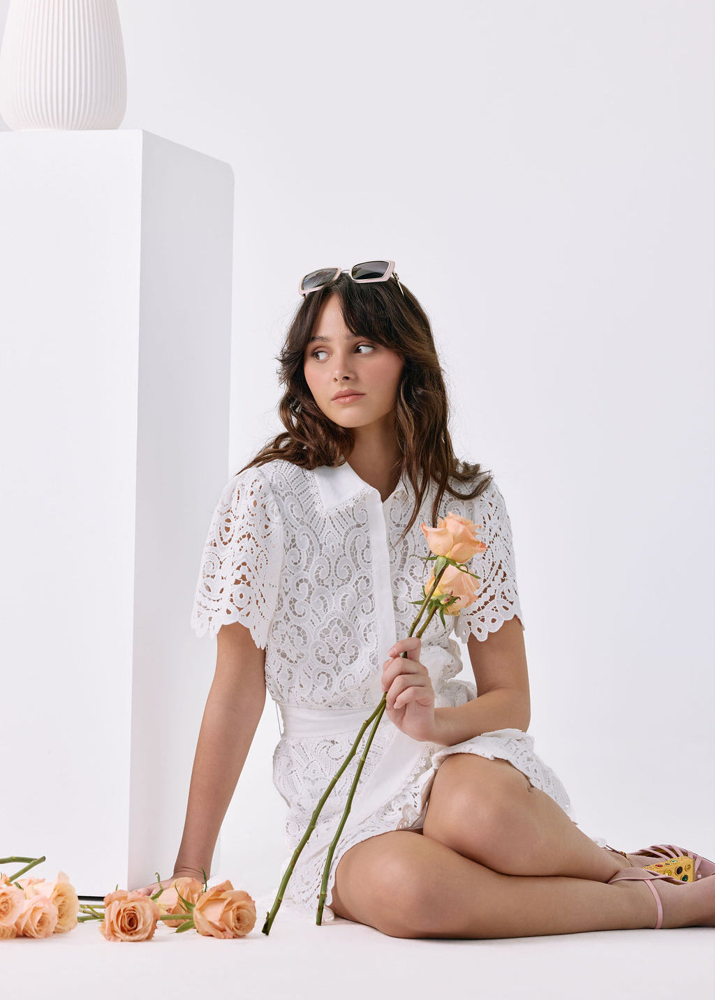 A woman sits cross-legged on a white floor beside a white pedestal, wearing a white lace short-sleeve top and white shorts. She holds peach roses, with more roses scattered on the floor.