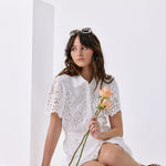 A woman sits cross-legged on a white floor beside a white pedestal, wearing a white lace short-sleeve top and white shorts. She holds peach roses, with more roses scattered on the floor.