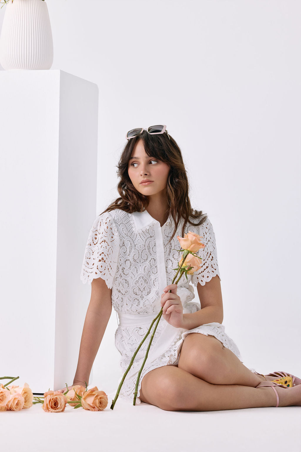 A woman sits cross-legged on a white floor beside a white pedestal, wearing a white lace short-sleeve top and white shorts. She holds peach roses, with more roses scattered on the floor.