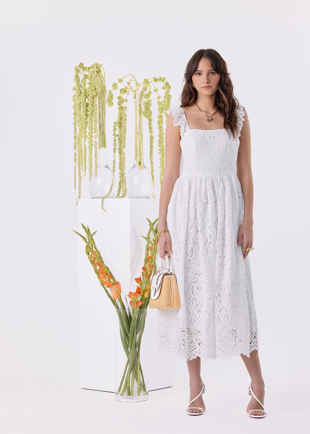 A woman modeling a white sleeveless midi dress with floral lace detailing, standing in a bright white studio next to a glass vase of orange flowers and a white pedestal with hanging greenery.