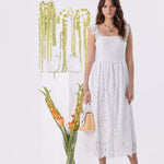 A woman modeling a white sleeveless midi dress with floral lace detailing, standing in a bright white studio next to a glass vase of orange flowers and a white pedestal with hanging greenery.