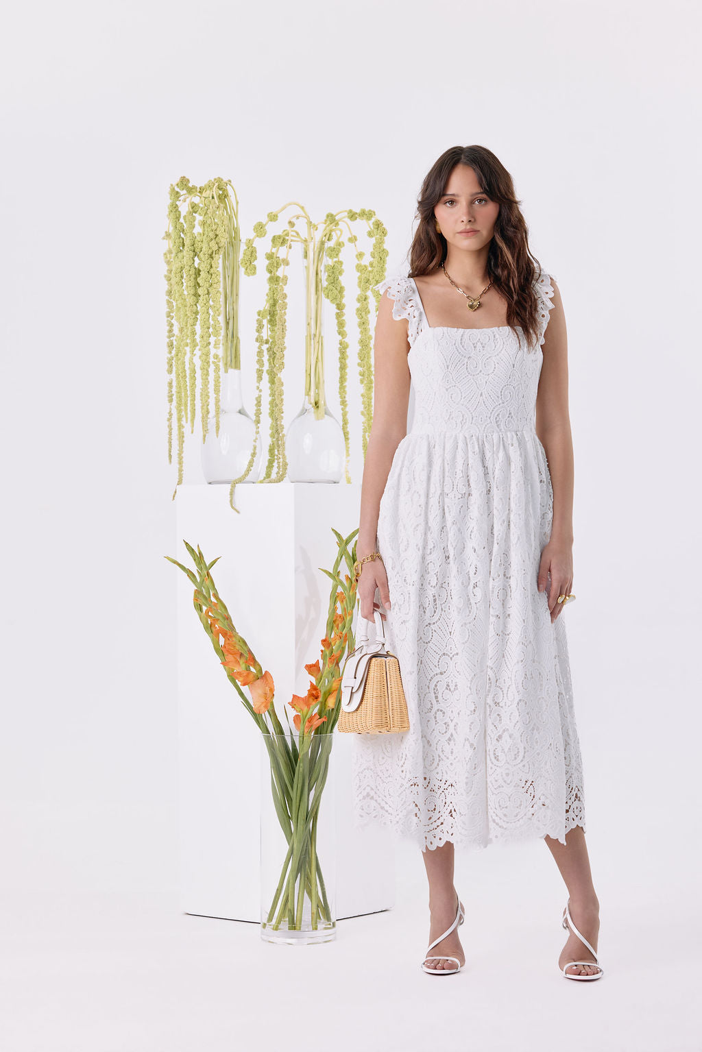 A woman modeling a white sleeveless midi dress with floral lace detailing, standing in a bright white studio next to a glass vase of orange flowers and a white pedestal with hanging greenery.