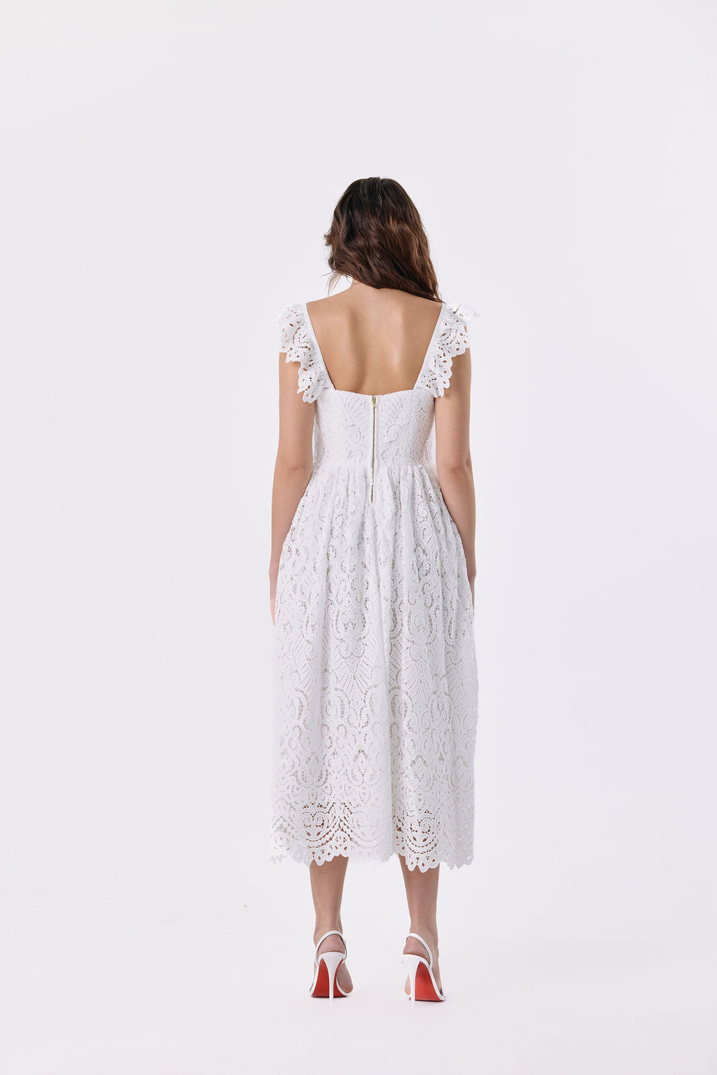 A woman viewed from the back wearing a white lace midi dress with ruffled cap sleeves and a square open back, paired with red high heels, against a plain white studio background.