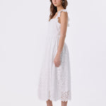 A woman modeling a white sleeveless lace midi dress with ruffled shoulder straps and a flared skirt, paired with white strappy high-heel sandals with red soles, against a plain white studio background.