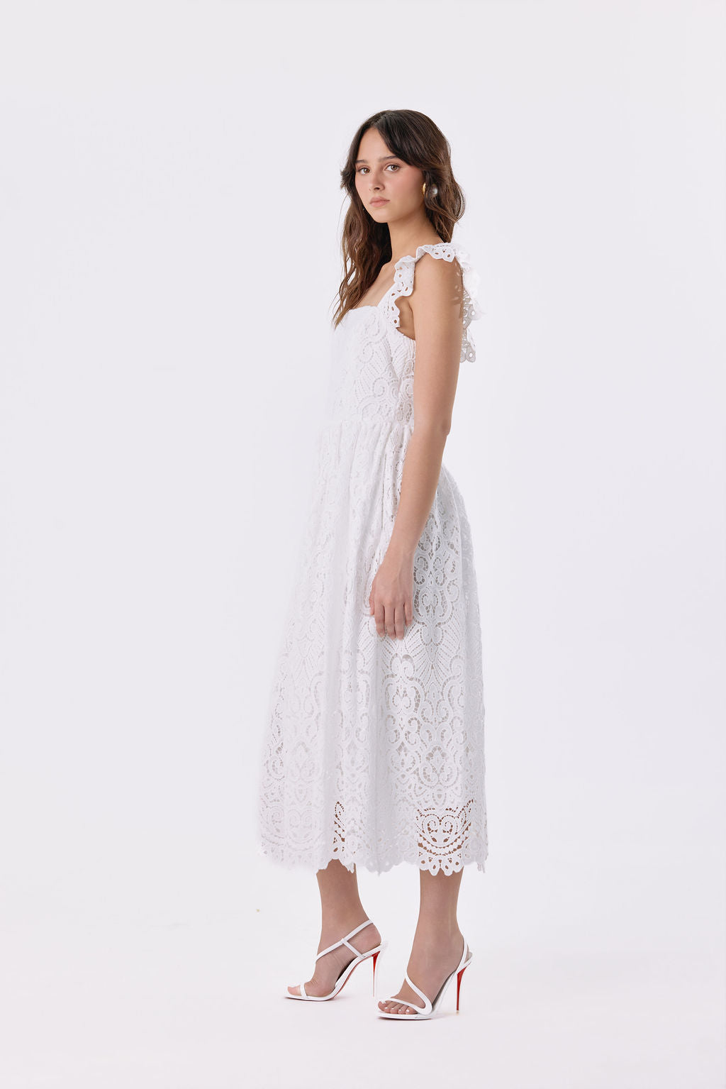 A woman modeling a white sleeveless lace midi dress with ruffled shoulder straps and a flared skirt, paired with white strappy high-heel sandals with red soles, against a plain white studio background.