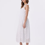 A woman modeling a white sleeveless lace midi dress with ruffled shoulder straps and a flared skirt, paired with white strappy high-heel sandals with red soles, against a plain white studio background.