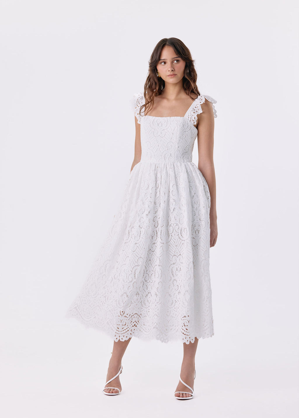 A woman modeling a white eyelet lace midi dress with ruffled shoulder straps, a square neckline, and a flared skirt, photographed against a seamless white studio background.