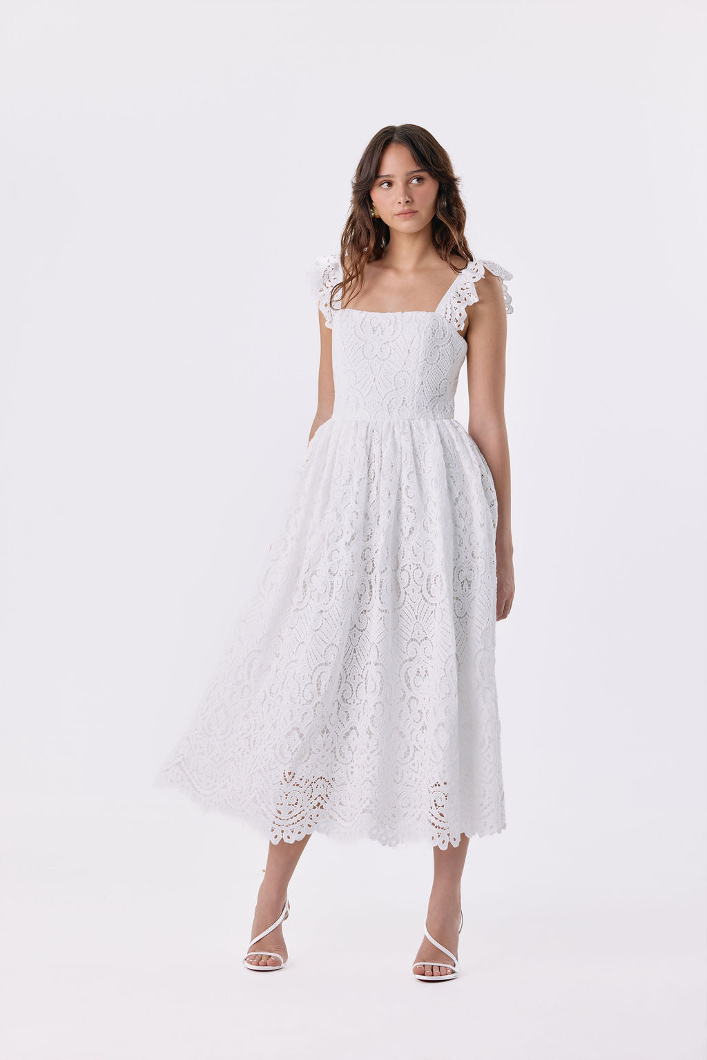 A woman modeling a white eyelet lace midi dress with ruffled shoulder straps, a square neckline, and a flared skirt, photographed against a seamless white studio background.