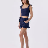 A model wearing a navy blue lace two-piece set consisting of a cropped top with ruffled shoulder details and a matching ruffled mini skirt, paired with beige high-heeled sandals, against a plain white studio background.