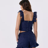 A woman shown from the back wearing a navy blue lace set with ruffled shoulder straps and a ruffled hem, photographed against a light gray studio background.
