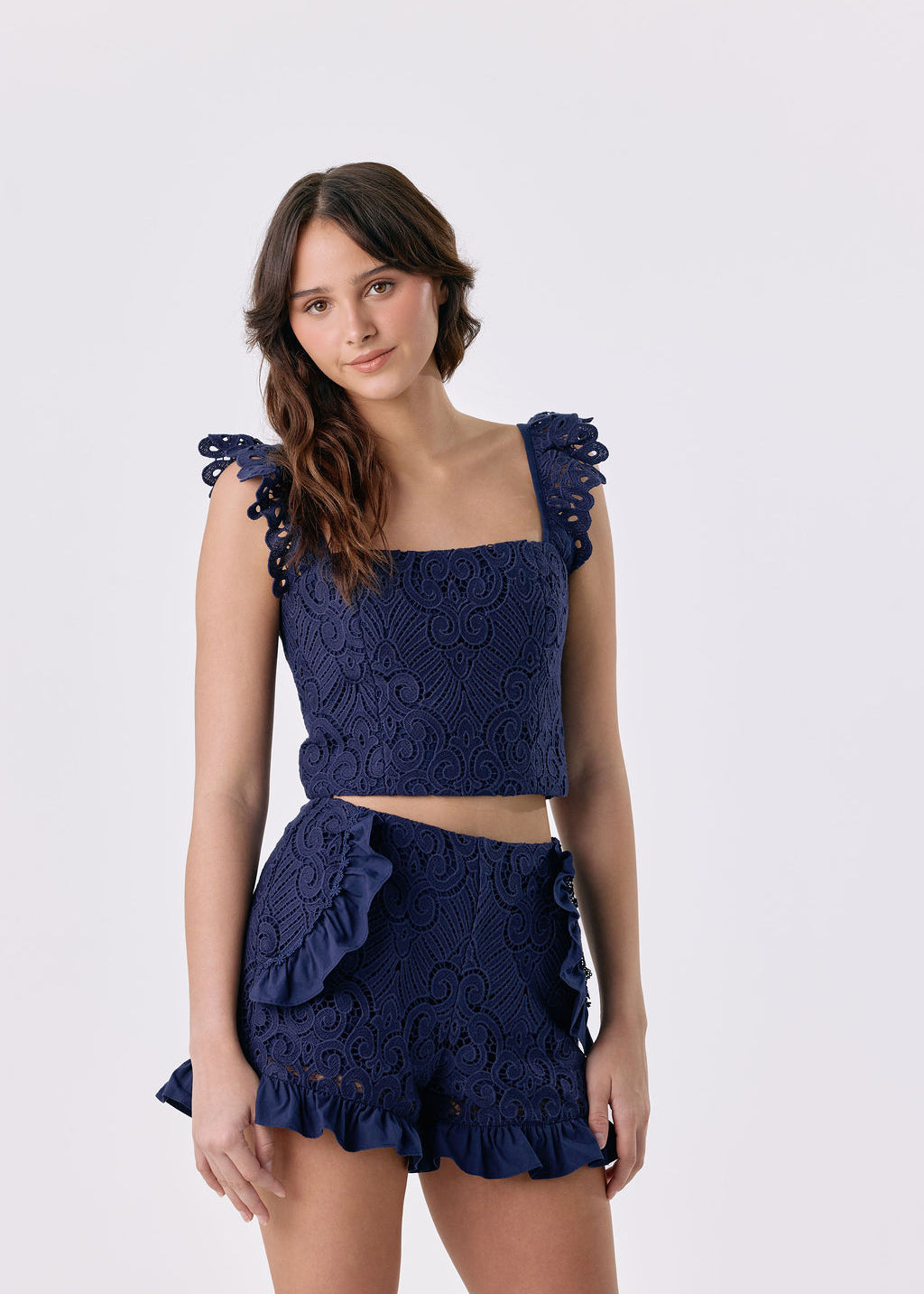 A woman modeling a navy blue two-piece set featuring a textured lace crop top with ruffled shoulder details and matching ruffled shorts, set against a plain light gray background.