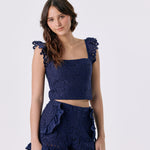 A woman modeling a navy blue two-piece set featuring a textured lace crop top with ruffled shoulder details and matching ruffled shorts, set against a plain light gray background.