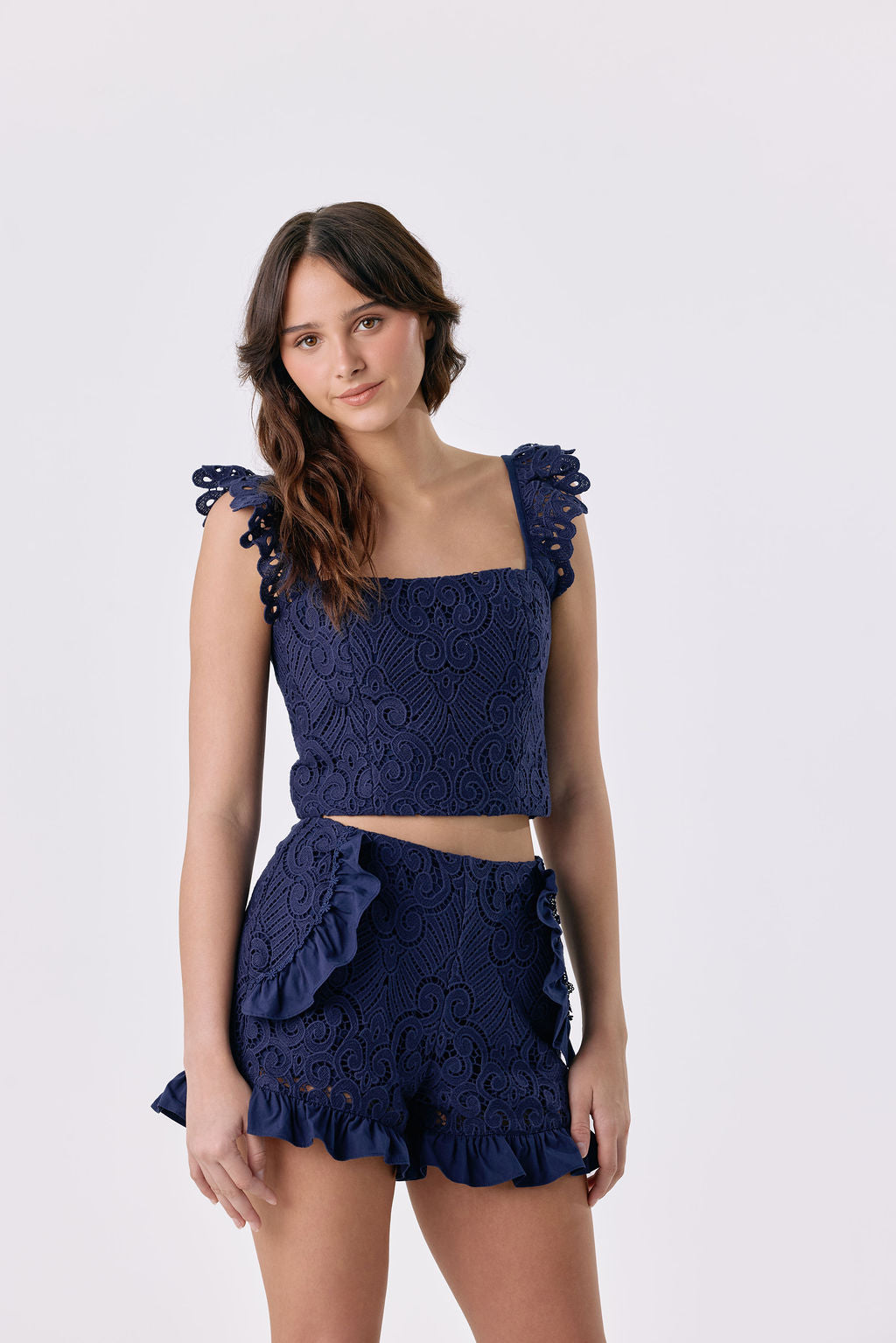 A woman modeling a navy blue two-piece set featuring a textured lace crop top with ruffled shoulder details and matching ruffled shorts, set against a plain light gray background.
