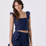 A woman modeling a navy blue two-piece set featuring a textured lace crop top with ruffled shoulder details and matching ruffled shorts, set against a plain light gray background.