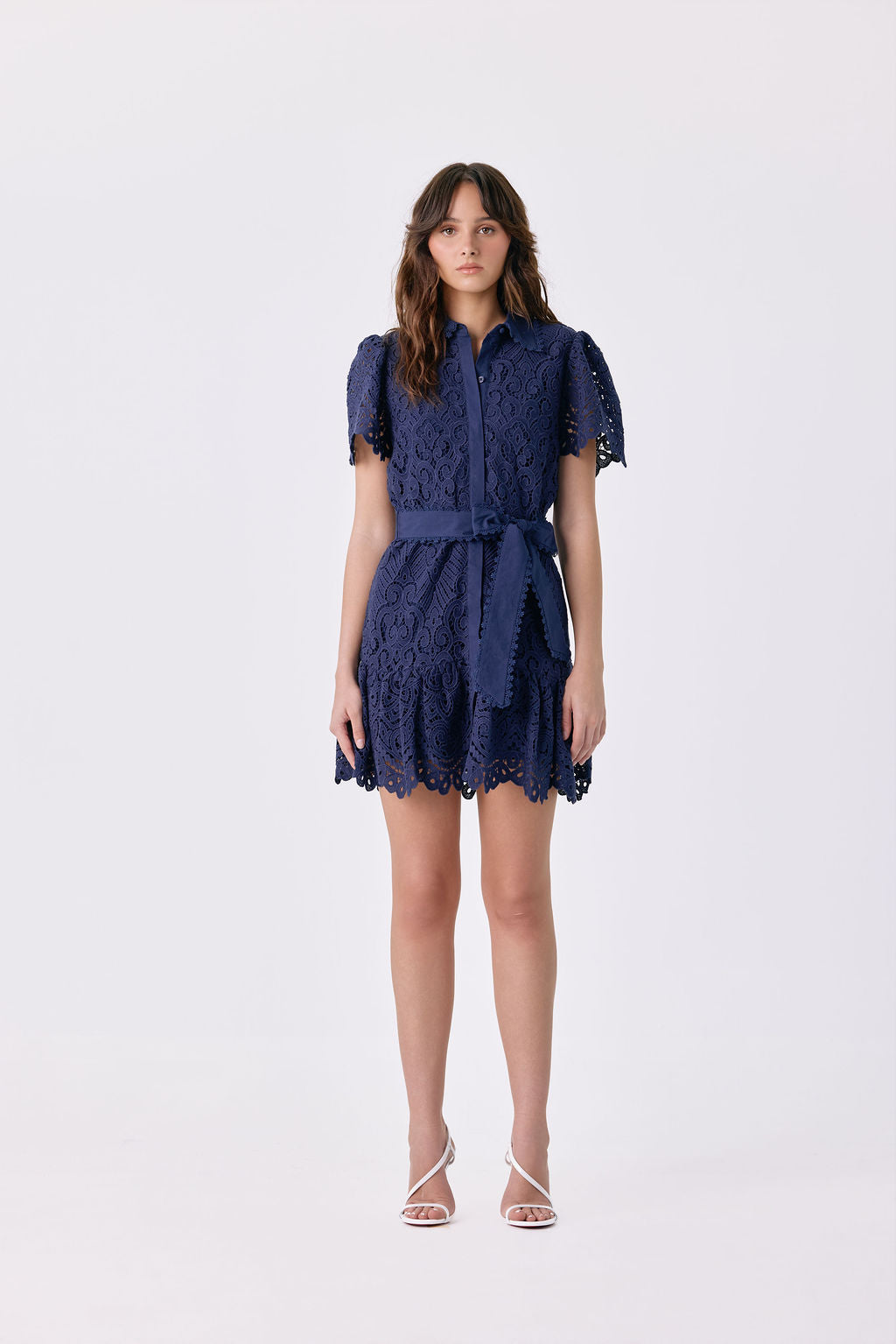 A woman modeling a navy blue lace mini dress with short puff sleeves, a button-up front, and a matching waist tie, photographed against a plain white studio background.
