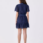 A woman viewed from the back wearing a navy blue lace mini dress with short puff sleeves and a ruffled hem, paired with white high heels with red soles, against a plain white studio background.