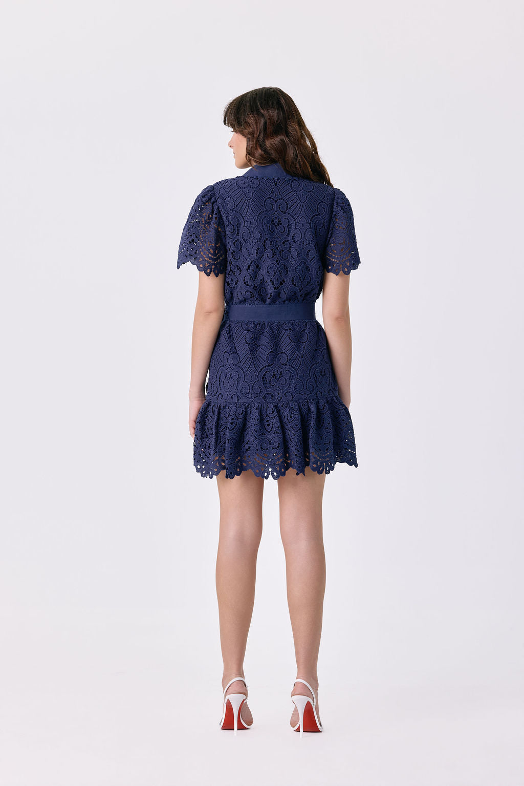 A woman viewed from the back wearing a navy blue lace mini dress with short puff sleeves and a ruffled hem, paired with white high heels with red soles, against a plain white studio background.