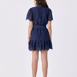 A woman viewed from the back wearing a navy blue lace mini dress with short puff sleeves and a ruffled hem, paired with white high heels with red soles, against a plain white studio background.