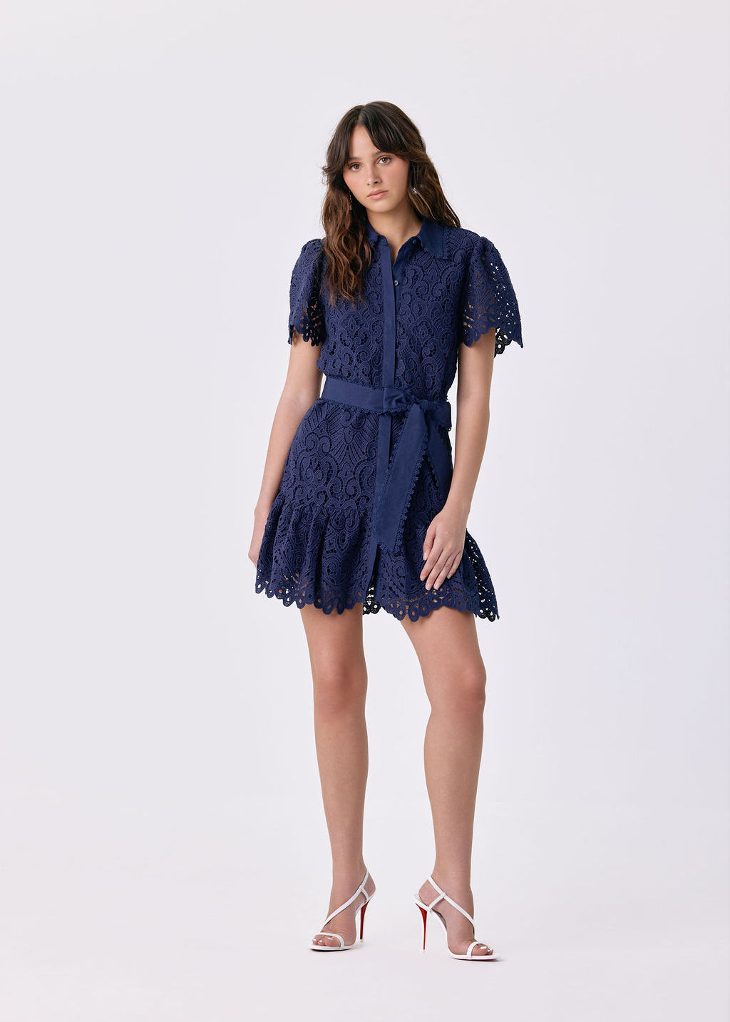 A woman modeling a short navy blue lace dress with short puff sleeves, a button-up front, and a matching waist tie, paired with white high-heeled sandals, against a light gray studio background.