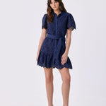 A woman modeling a short navy blue lace dress with short puff sleeves, a button-up front, and a matching waist tie, paired with white high-heeled sandals, against a light gray studio background.