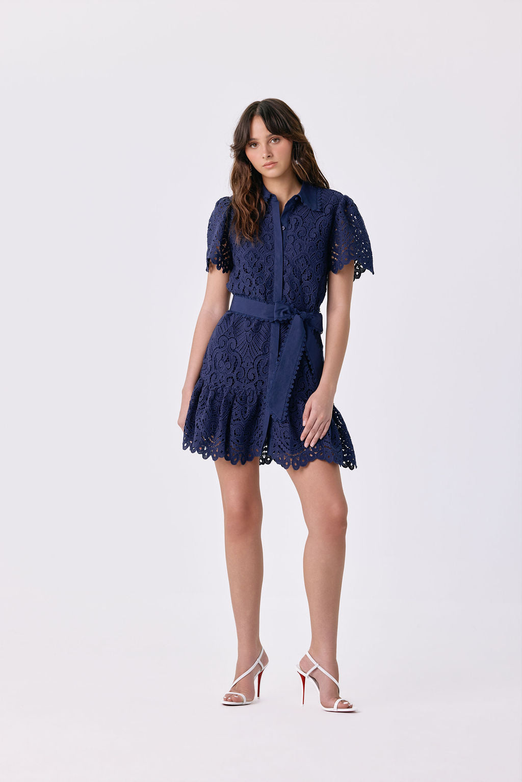 A woman modeling a short navy blue lace dress with short puff sleeves, a button-up front, and a matching waist tie, paired with white high-heeled sandals, against a light gray studio background.