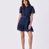 A woman modeling a short navy blue lace dress with short puff sleeves, a button-up front, and a matching waist tie, paired with white high-heeled sandals, against a light gray studio background.
