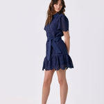 A woman modeling a short navy blue lace dress with short sleeves, a tie waist, and a ruffled hem, paired with white strappy high-heeled sandals, against a plain white studio background.