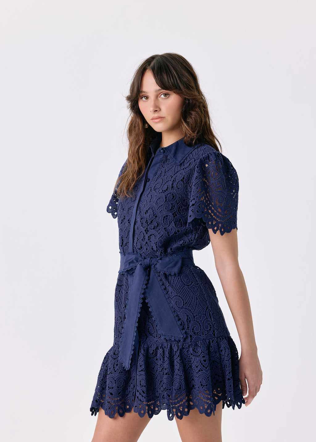 A woman modeling a navy blue lace mini dress with short puff sleeves, a tie waist, and a ruffled hem against a light gray studio background.