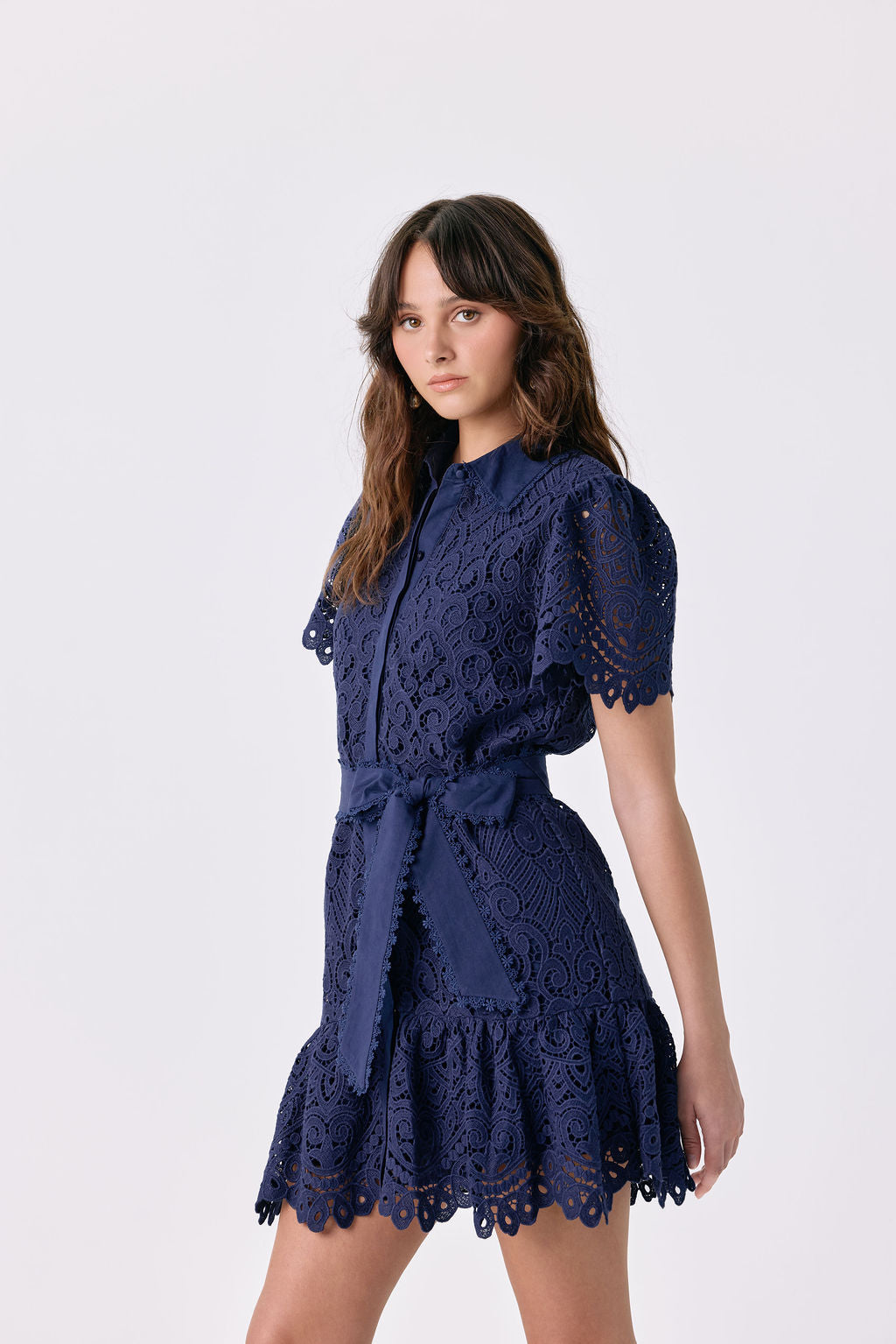 A woman modeling a navy blue lace mini dress with short puff sleeves, a tie waist, and a ruffled hem against a light gray studio background.