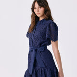 A woman modeling a navy blue lace mini dress with short puff sleeves, a tie waist, and a ruffled hem against a light gray studio background.