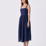 A woman modeling a navy blue lace midi dress with thin straps and a flared skirt, paired with white heeled sandals, against a plain white studio background.