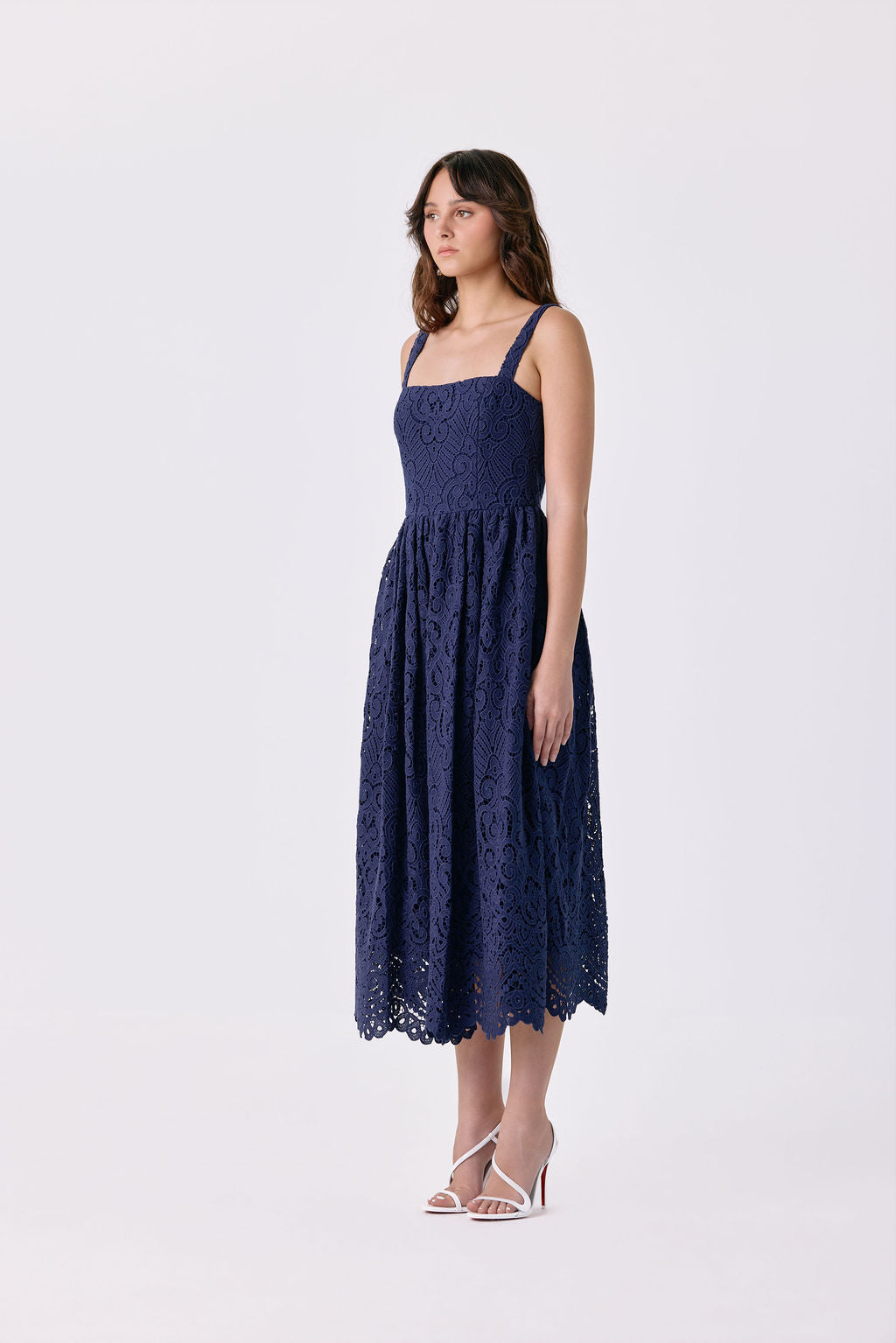 A woman modeling a navy blue lace midi dress with thin straps and a flared skirt, paired with white heeled sandals, against a plain white studio background.