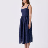 A woman modeling a navy blue lace midi dress with thin straps and a flared skirt, paired with white heeled sandals, against a plain white studio background.