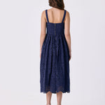 A woman viewed from the back wearing a sleeveless navy blue lace midi dress with thin shoulder straps and a gathered waist, paired with red high-heeled sandals, against a plain light gray studio background.