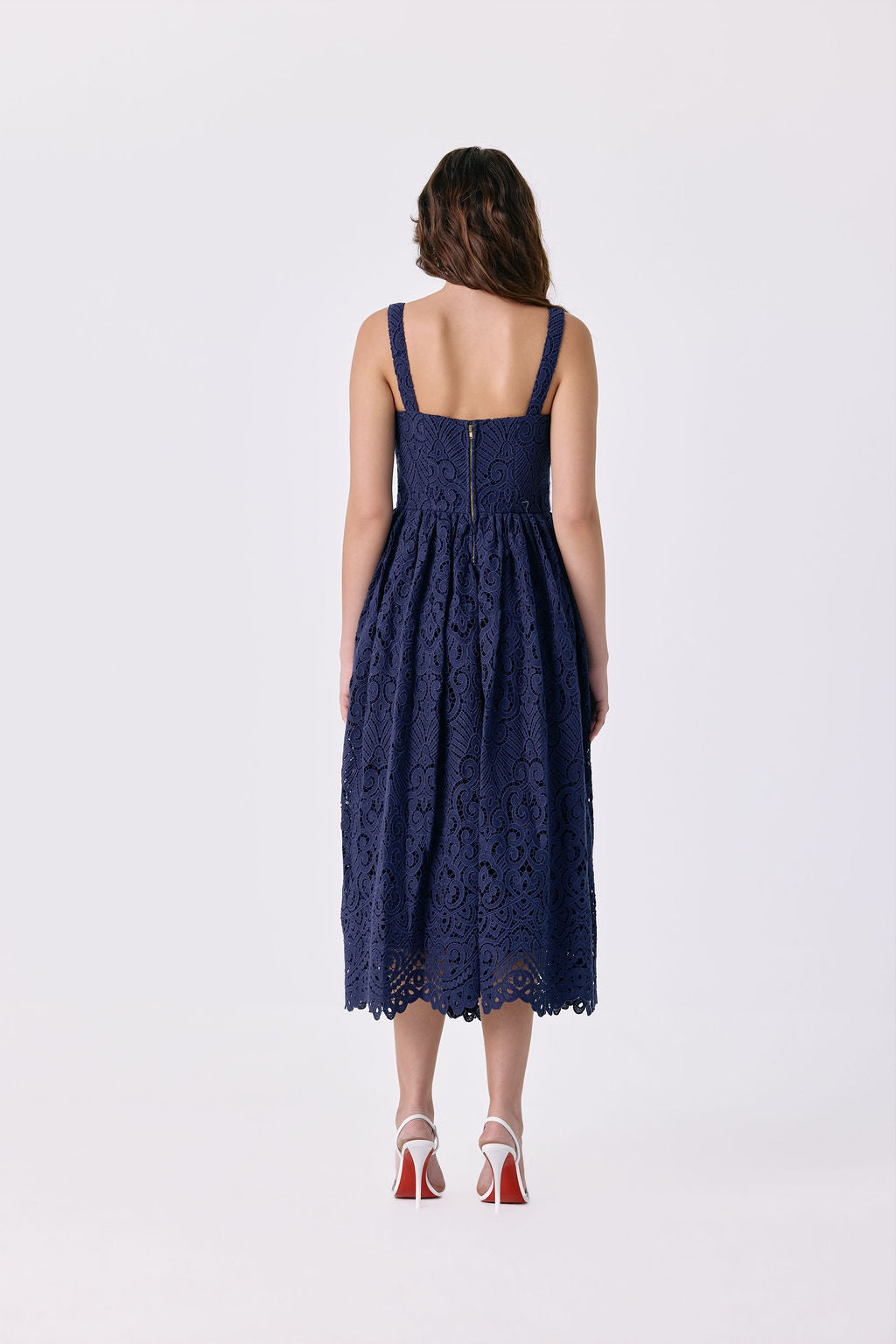 A woman viewed from the back wearing a sleeveless navy blue lace midi dress with thin shoulder straps and a gathered waist, paired with red high-heeled sandals, against a plain light gray studio background.